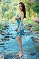 A woman in a blue dress standing on a rock by a river.